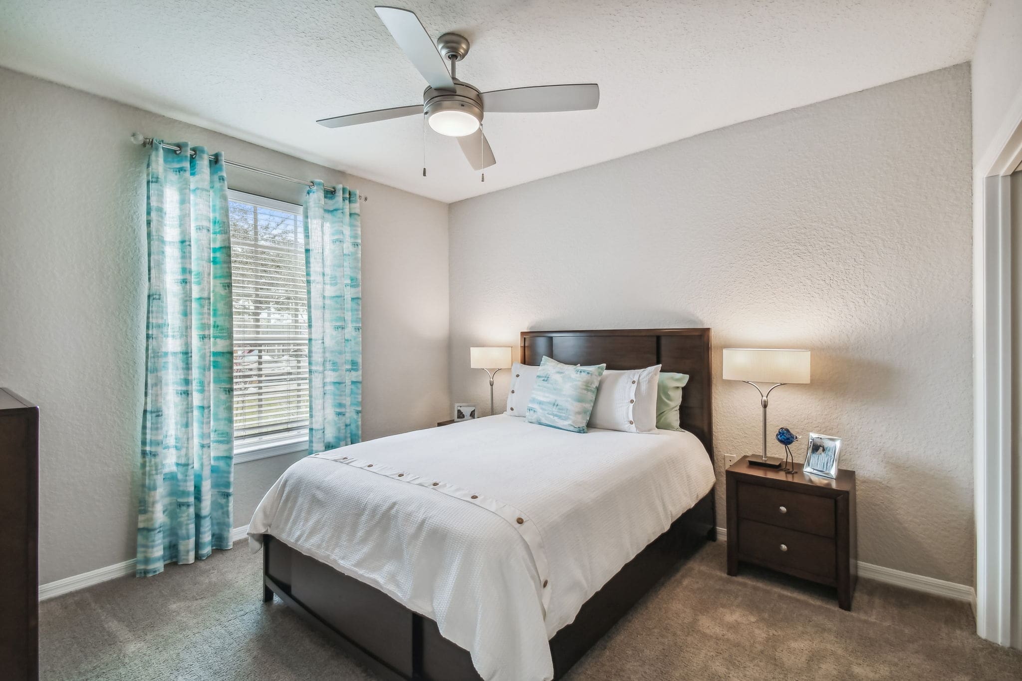 Brandon, FL, Apartments Near I-75 Interchange - 2211 Grand Isle - Bedroom With White Bedding, Wood Headboard, Nightstands, Lamps, Ceiling Fan, and Large Window With Blinds and Curtains